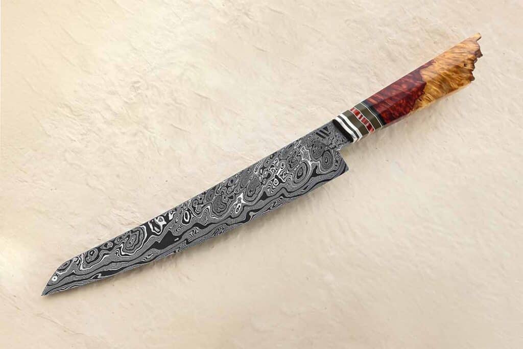 For a great example of a go-mai (five layer) damascus blade with the visual “pop” of nickel, Robert Wayman provides the fireworks via his sujihiki.  Damascus outer layers and a damascus core work together with the handle of live edge amboyna burl to make a stunning knife. Overall length: 16 inches. (SharpByCoop image)