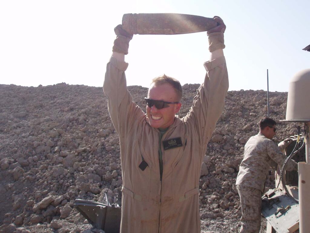 USMC Staff Sergeant Les George holds an artillery shell aloft somewhere in Iraq in the early 2000s.