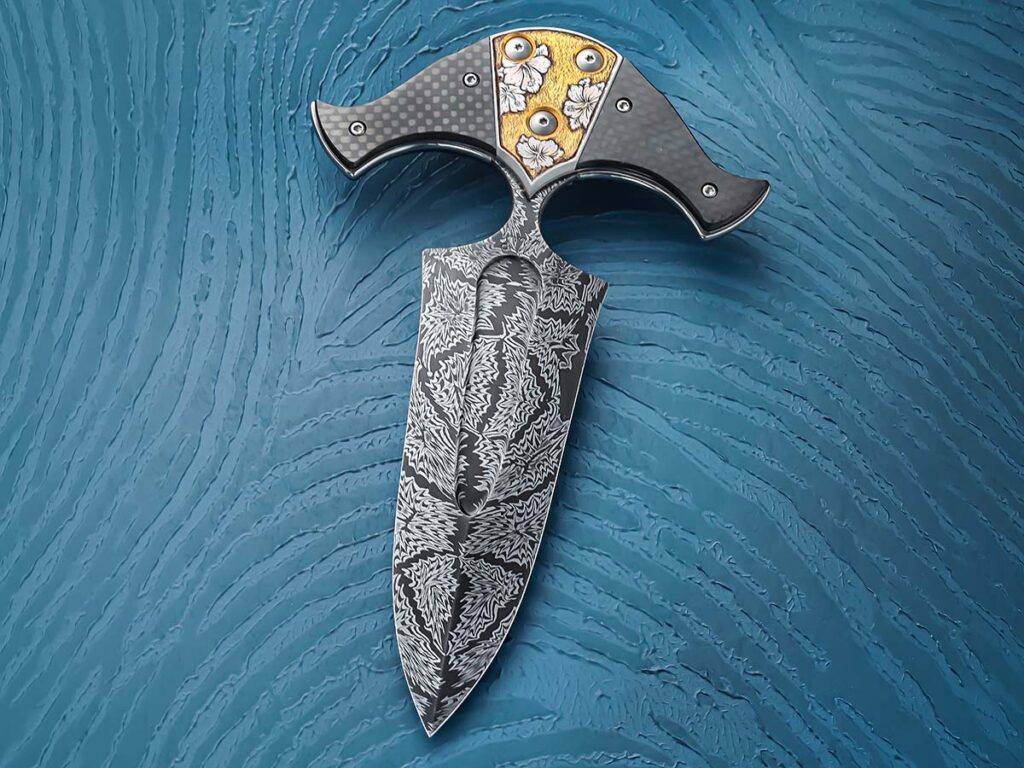 A bold mix of materials, Andrew Meers utilizes carbon fiber, stainless steel,  gold and a splintered mosaic damascus to bring-together this 9-inch push dagger. The centerpiece of the knife is its elegant Rose of Sharron engraving with the gold backing. (SharpByCoop image)