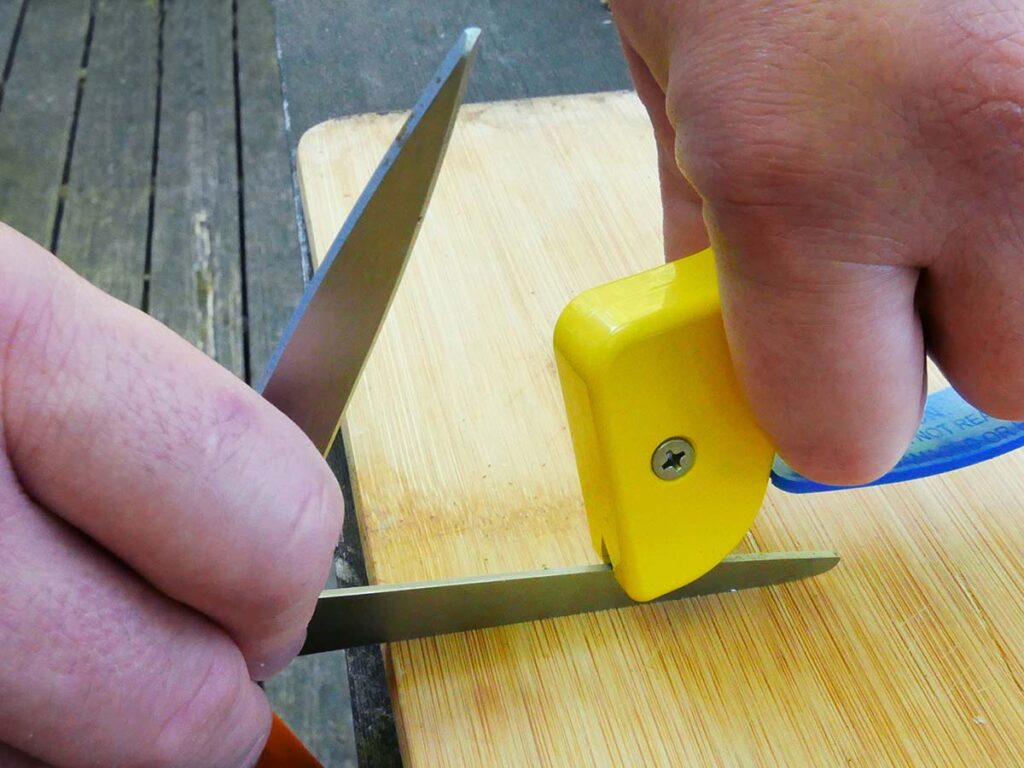 The AccuSharp ShearSharp Scissors Sharpener puts an edge on the blade bevels one side at a time. Here the blades are opened with one side resting on the flat work surface having the blade bevel sharpened. Note the other blade is off the surface. Once the first blade is done, the scissors are flipped and the second blade is sharpened in the same manner. (Covert image)